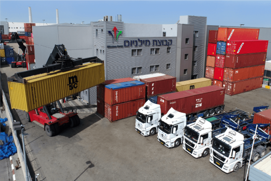 בתמונה רואים את שירותי הובלה ורכבת של קבוצת מילניום מסוף אשדוד מוצג בתמונה שבו משאיות ההובלה של קבוצת מילניום חונות בשורה עם מכולות מועמסות עליהן והלוגו של מילניום נמצא על הבניין מאחוריהן