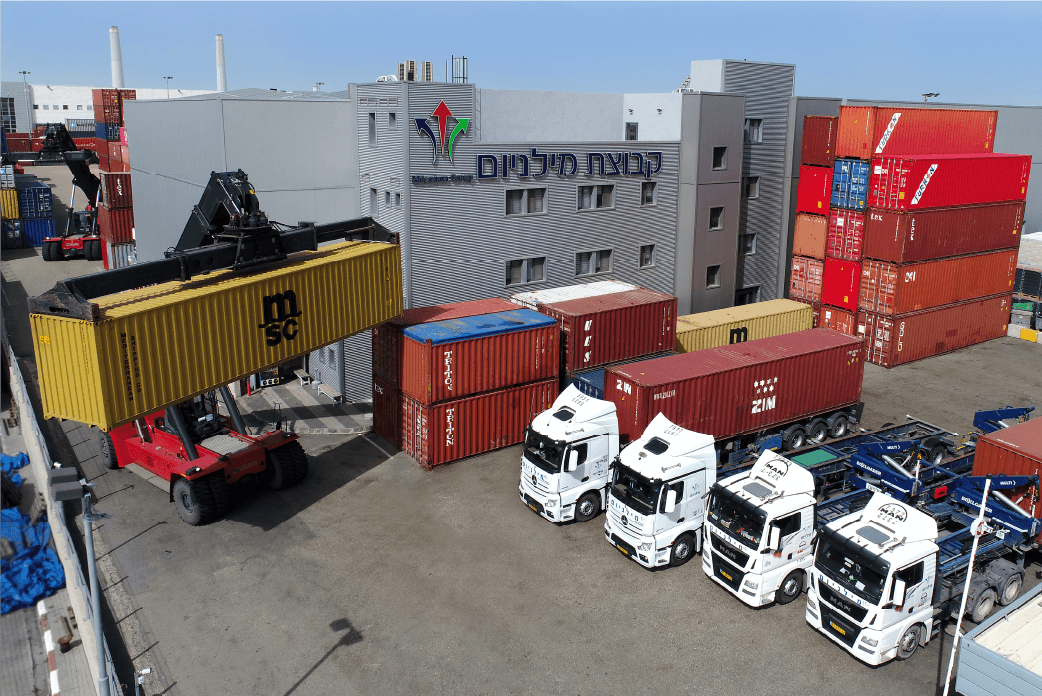 בתמונה רואים את שירותי הובלה ורכבת של קבוצת מילניום מסוף אשדוד מוצג בתמונה שבו משאיות ההובלה של קבוצת מילניום חונות בשורה עם מכולות מועמסות עליהן והלוגו של מילניום נמצא על הבניין מאחוריהן
